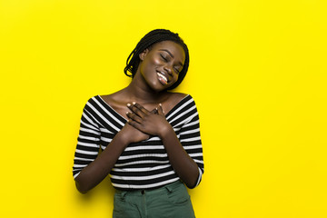 Wall Mural - Headshot of pleased touched young African American woman smiles gently, feels pleased, wears striped top and headwear, isolated over yellow background. People, happiness and emotions concept