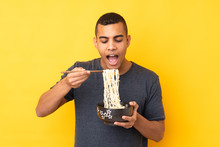 Young African American Man Over Isolated Yellow Background Holding A Bowl Of Noodles With Chopsticks And Eating It
