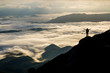 © bilanol - Wide mountain panorama. Small silhouette of tourist with backpack on rocky mountain slope with raised hands over valley covered with white puffy clouds. Beauty of nature, tourism and traveling concept