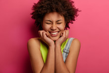 Head Shot Of Positive Curly Haired Woman Keeps Hands Under Chin, Closes Eyes And Smiles Broadly, Enjoys Nice Time In Good Company, Dressed In Fashionable Clothes, Isolated On Pink Background