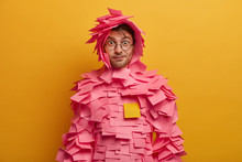 Indoor Shot Of Surprised Funny Man Looks Through Transparent Glasses, Wears Paper Costume Has Direct Gaze At Camera Isolated Over Yellow Background. Wondered Guy Dressed In Outfit Made Of Sticky Notes
