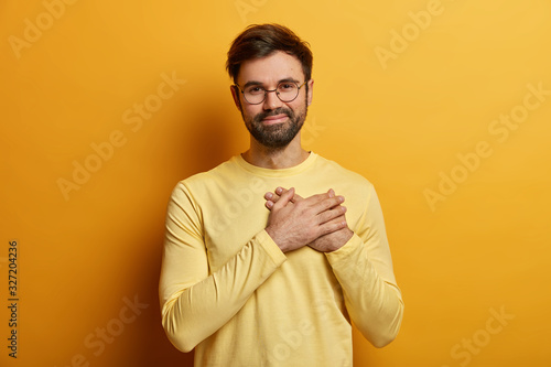 Fotomural Handsome bearded guy keeps hands on heart, expresses sincere emotions, appreciates help and heartwarming words, stands thankful, wears casual yellow jumper, poses indoor