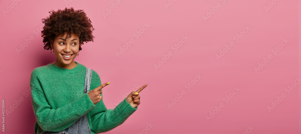 Positive ethnic woman points at product, suggests check it out, indicates right with happy smile, rejoices nice advertisement, wears green sweater, poses against pink background. Promo concept - obrazy, fototapety, plakaty 