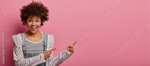Fotografía Lively excited curly haired woman points fingers aside, being fully pleased with
