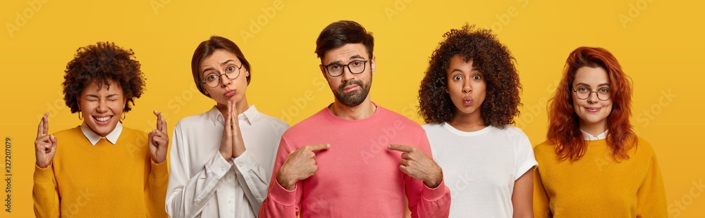 Unimpressed bearded man points at himself, four women stand near him, pray and beg for something, have glad expressions, wear spectacles, isolated over yellow background. Body language, emotions - obrazy, fototapety, plakaty 