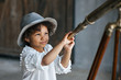 © Olena - Close-up portrait of a cute little african american kid girl in a white dress with a leather belt and a gray hat looks through the telescope. Studio shooting.