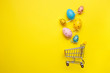 Shopping trolley with Easter eggs and copy space on yellow background. Easter shopping and sale concept. Flat lay, top view