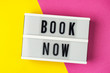 Book now - text on a display lightbox on yellow and pink background.