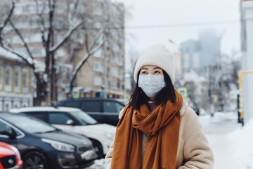 Wall Mural - the girl next to the cars and against the background of high-rise buildings looks at the camera in a protective medical mask. The concept of coronavirus in everyday life