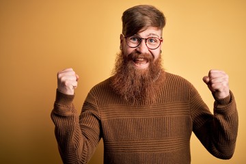 Handsome Irish redhead man with beard wearing glasses and winter sweater over yellow background celebrating surprised and amazed for success with arms raised and open eyes. Winner concept.