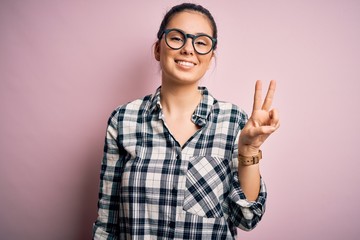 Young beautiful brunette woman wearing casual shirt and glasses over pink background smiling with happy face winking at the camera doing victory sign. Number two.