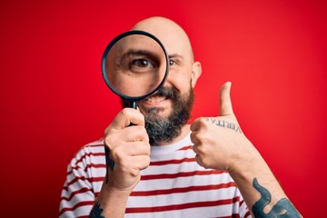 Sticker - Handsome detective bald man with beard using magnifying glass over red background happy with big smile doing ok sign, thumb up with fingers, excellent sign