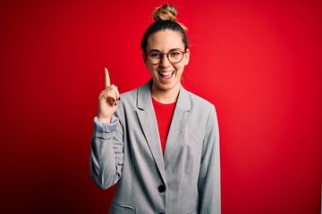 Young beautiful blonde businesswoman with blue eyes wearing glasses and jacket pointing finger up with successful idea. Exited and happy. Number one.