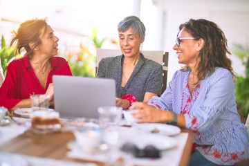 Wall Mural - Meeting of middle age women having lunch and drinking coffee. Mature friends smiling happy using laptop at home on a sunny day