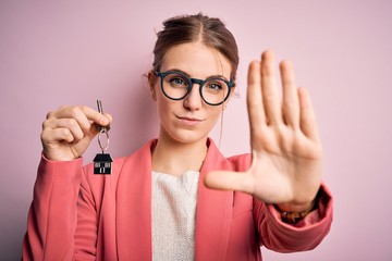 Wall Mural - Young beautiful redhead house agent woman holding home key over pink bakcground with open hand doing stop sign with serious and confident expression, defense gesture