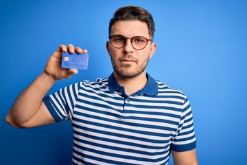 Young man with blue eyes wearing glasses and holding credit card over blue background with a confident expression on smart face thinking serious