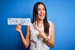 © Krakenimages.com - Young beautiful brunette woman holding paper with tax day message over blue background very happy pointing with hand and finger