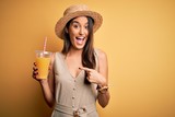 Young beautiful woman on vacation wearing summer hat drinking healthy orange juice very happy pointing with hand and finger