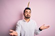 Young handsome man with beard wearing funny unicorn diadem over pink background clueless and confused expression with arms and hands raised. Doubt concept.