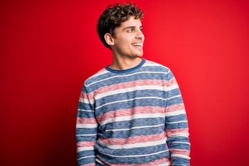 Young blond handsome man with curly hair wearing striped sweater over red background looking away to side with smile on face, natural expression. Laughing confident.