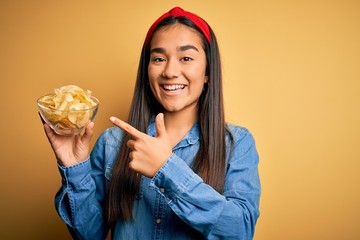 Canvas Print - Young beautiful asian woman holding bowl with chips potatoes over isolated yellow background very happy pointing with hand and finger