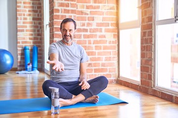 Canvas Print - Middle age handsome sportman sitting on mat doing stretching yoga exercise at gym smiling cheerful offering palm hand giving assistance and acceptance.