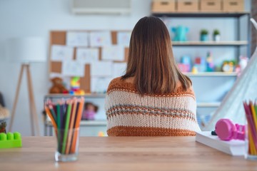 Poster - Young beautiful teacher woman wearing sweater and glasses sitting on desk at kindergarten standing backwards looking away with crossed arms