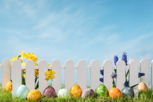 Spring Grass And Wooden Fence With Easter Eggs And Flower On Cloudy Sky