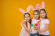 © producer - happy cute little child girls with pink bunny ears holding painted Easter eggs on studio yellow background. Easter day