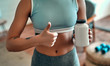 © Valerii Apetroaiei - Cropped image of an athletic woman in sportswear holds a jar of protein in her hand and showing thumbs up gesture at home in the living room.