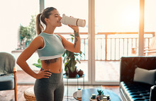 Athletic Woman In Sportswear Drinking A Protein Shake Or A Bottle Of Water At Home In The Living Room. Sport And Recreation Concept.