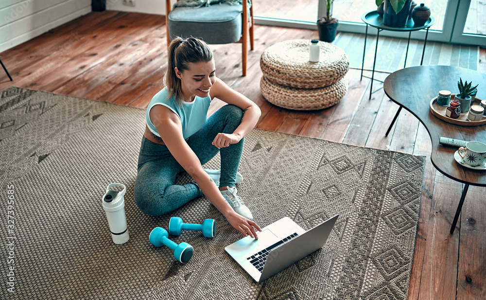A sporty woman in sportswear is sitting on the floor with dumbbells and a protein shake or a bottle of water and is using a laptop at home in the living room. Sport and recreation concept. - obrazy, fototapety, plakaty 