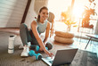 © Valerii Apetroaiei - A sporty woman in sportswear is sitting on the floor with dumbbells and a protein shake or a bottle of water and is using a laptop at home in the living room. Sport and recreation concept.