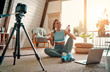 © Valerii Apetroaiei - Woman blogger in sportswear sitting on the floor with dumbbells and a laptop and showing a jar of sports nutrition proteins to the camera at home in the living room. Sport and recreation concept.