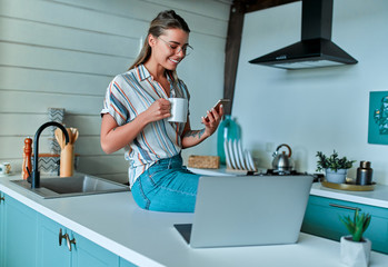 A cheerful young woman in casual clothes and glasses sits on a countertop with a cup of coffee and a phone while working on a laptop at home in her kitchen. Freelance work at home.