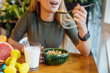 Cropped image, sport woman eating corn flakes cereal with milk, fresh oranges and dumbbells at the kitchen table