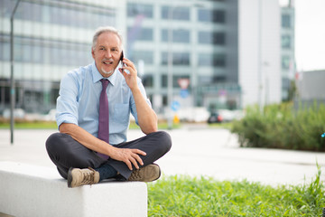 Wall Mural - Businessman using his cellphone outdoor while sitting on a bench in front of his office