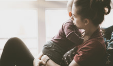 Concerned Woman Holding And Hugging A Small Child. Mom From The Generation Of Milenials Sits By The Window And Holds The Son. Slight Noise And Shallow Depth Of Field.