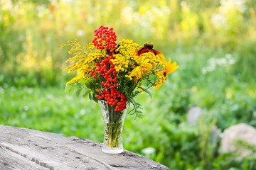 Handmade bouquet of wildflowers. Rowan berries on branch, goldenrog plant and tansy flowers. Floral composition in transparent vase on summer nature background outdoors.