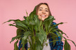 © khosrork - Closeup of funny housewife peeping from bush of green plant and looking at camera with pleased smile, holding big flowerpot, loves gardening and nature. indoor studio shot isolated on pink background