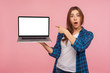 Portrait of amazed charming girl in checkered shirt showing blank laptop screen with shocked expression, pointing mock up, copy space for internet advertising. studio shot isolated on pink background