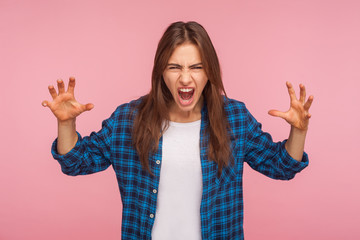 Portrait of enraged girl in checkered shirt with furious crazy grimace raising hands and shouting in hatred, anger management problem, irritation. indoor studio shot isolated on pink background