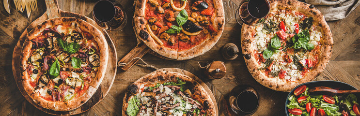 Pizza party dinner. Flat-lay of various kinds of Italian pizza, salad and red wine in glasses over rustic wooden table, top view, wide composition. Fast food lunch, celebration, gathering concept