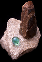 Top Down View Of A Vibrant Teal Green Glass Marble Reflecting Its Light On The Textured Surface Of A White Stone With Brown Mining Rock Vertical Besides It