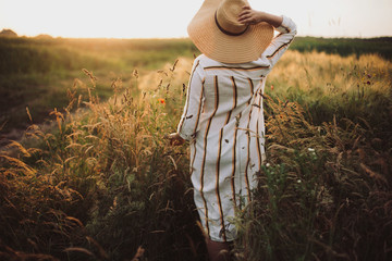Wall Mural - Woman in rustic dress and hat walking in wildflowers and herbs in sunset golden light in summer meadow. Atmospheric authentic moment. Stylish girl enjoying evening in countryside.