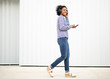 © mimagephotos - Full length portrait young african american woman walking listening to music with cellphone and headphones by white wall