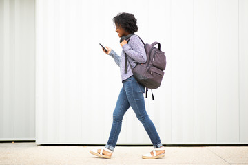 full length side young african american woman walking with mobile phone and bag by white wall