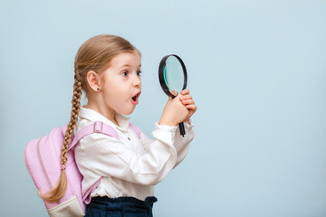 Cheerful young blonde baby girl looks through a magnifying glass. isolated on a blue background. Looks at the world with surprise.