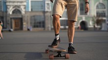 The Skater's Legs Begin To Move On A Skateboard In The Fresh Air. One Leg Stands On The Asphalt, The Other On The Longboard. Sunny Blurred Background.
