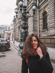 Portrait of a beautiful brunette girl with long hair in a coat and a bright scarf, stands against the background of a beautiful old building with columns, a lantern in St. Petersburg